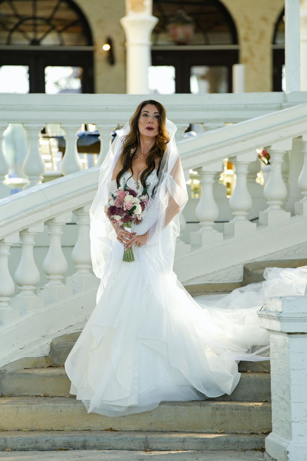 Bride in white mermaid gown and veil holds bouquet on elegant white marble staircase outdoors.