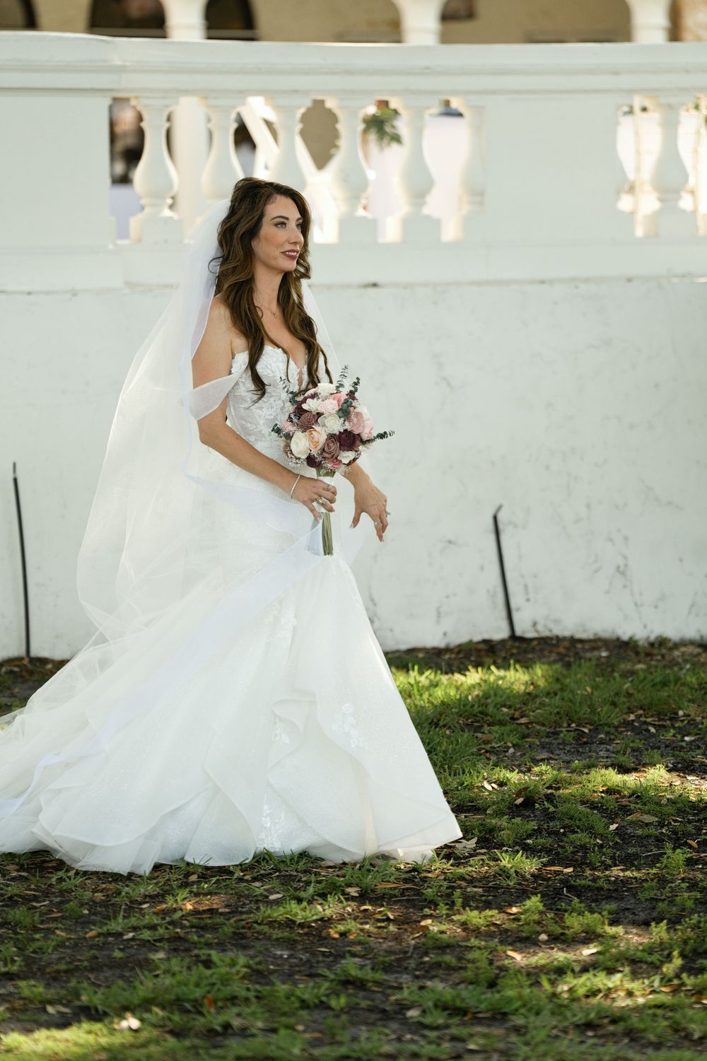 Bride in white off-shoulder mermaid gown holding floral bouquet, smiling outdoors near white balustrade.