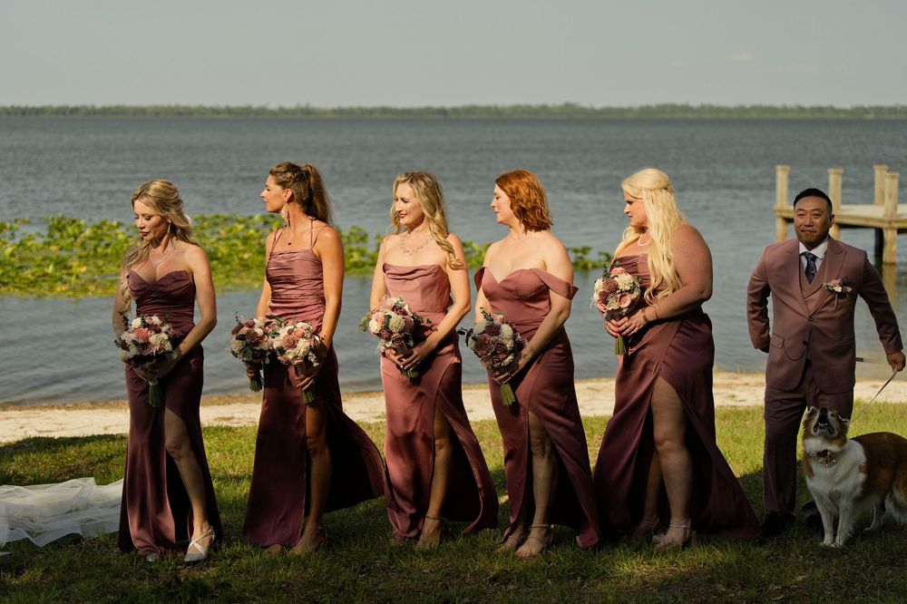 Five bridesmaids in mauve gowns holding bouquets stand by a waterfront with a groomsman and dog on a sunny day.