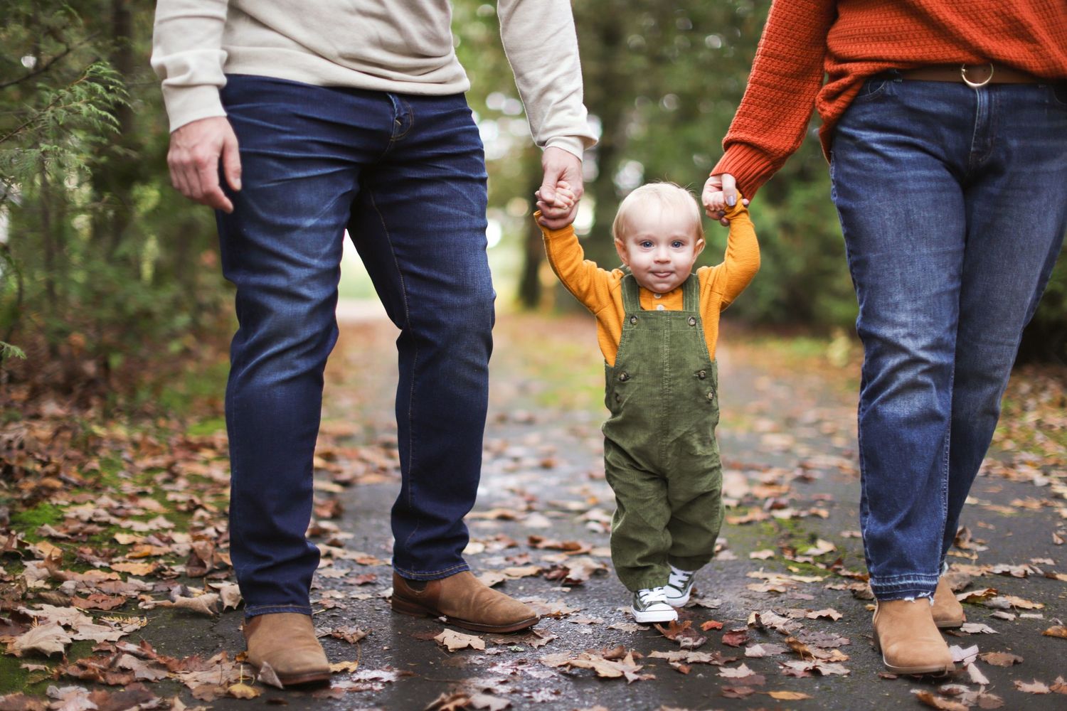 Parents holding toddler's hands while walking on a leaf-covered autumn path in a forest park.
