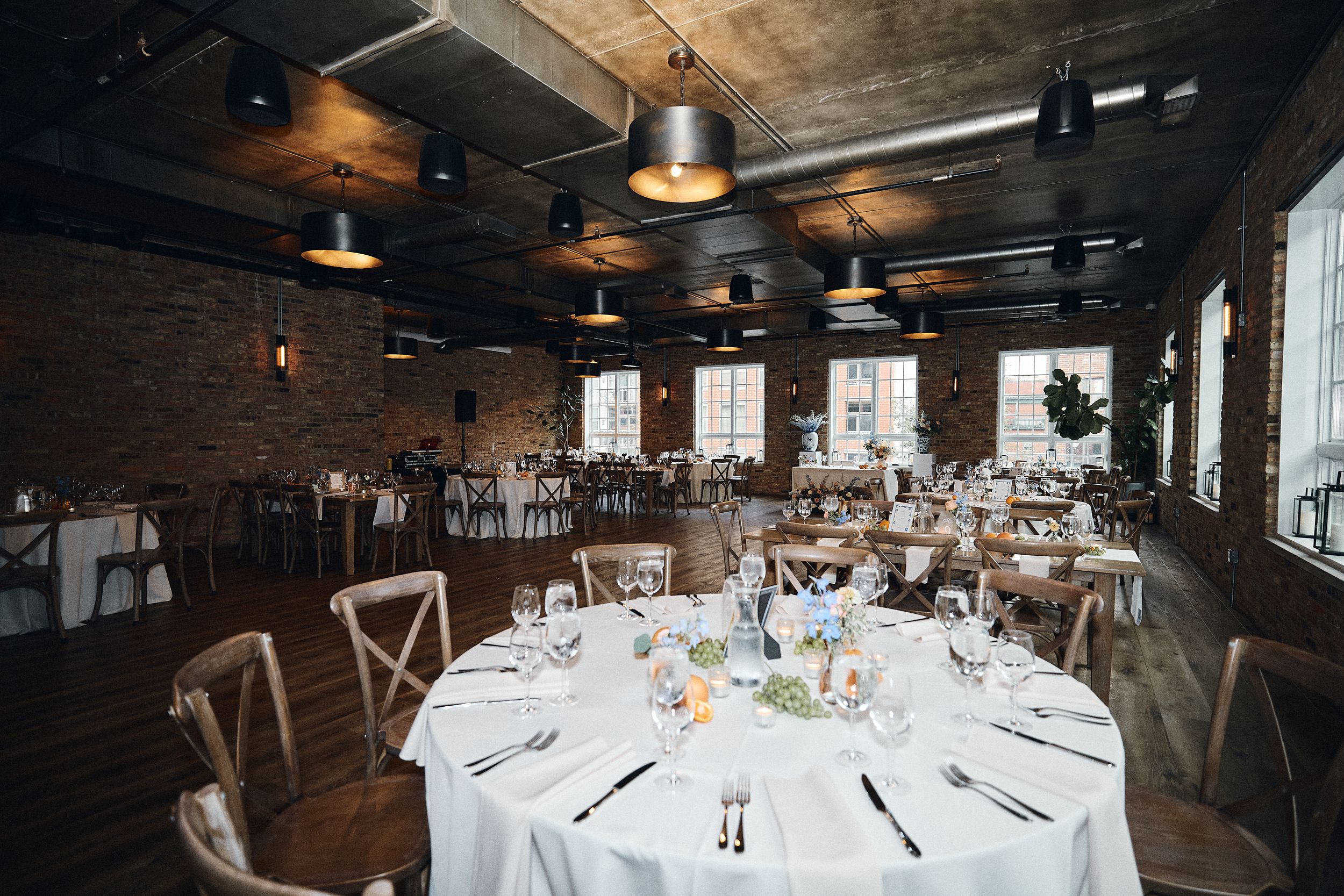 Elegant wedding reception hall with round tables, white linens, cross-back chairs, exposed brick walls, and industrial pendant lights.