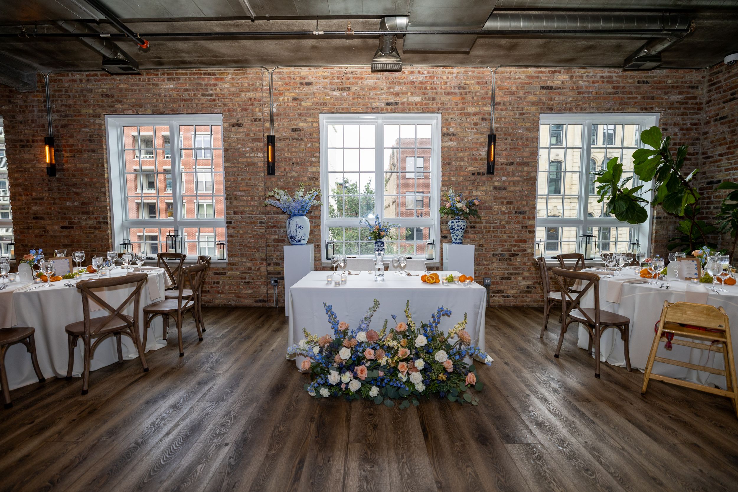 Elegant wedding reception venue with brick walls, white draped tables, floral centerpieces, and wood floors.