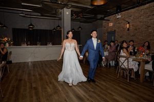 Bride in white gown and groom in blue suit hold hands entering wedding reception on hardwood dance floor.