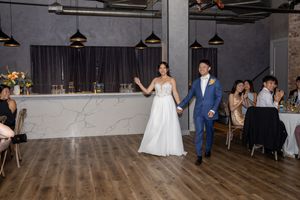 Bride in white gown and groom in blue suit hold hands entering their wedding reception in an elegant industrial venue.