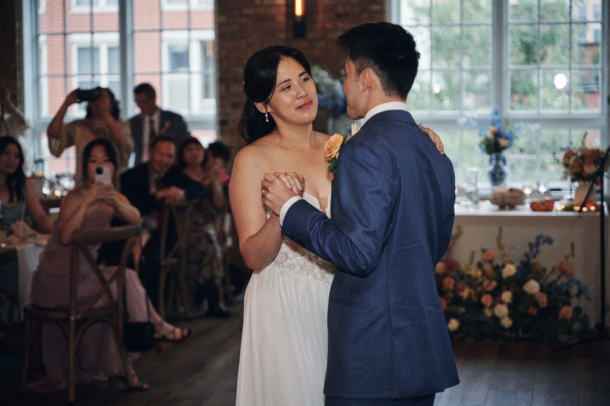 Bride and groom share their first dance at wedding reception, guests watching and photographing the romantic moment.