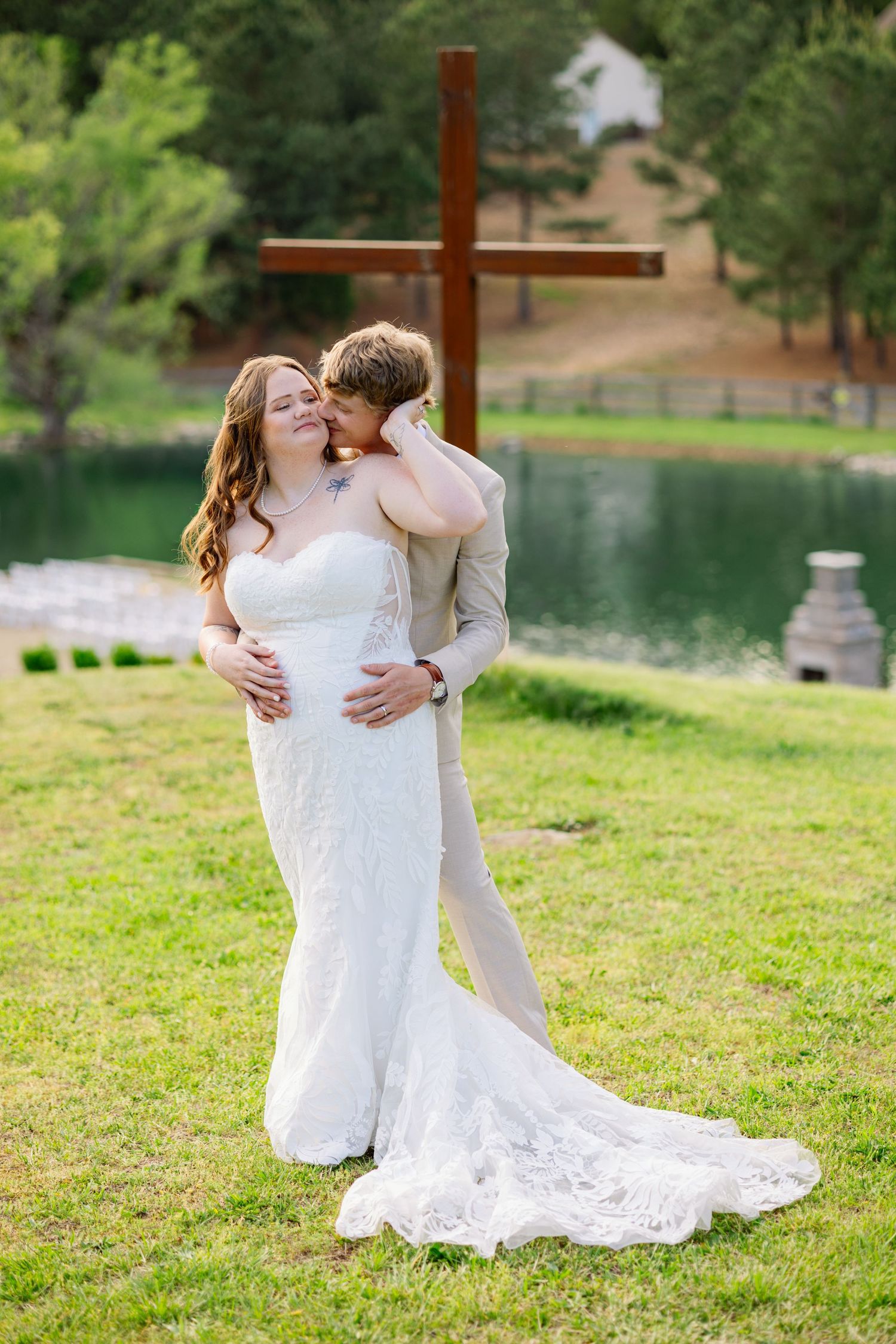 Bride and groom embrace outdoors near a wooden cross and pond, bride in lace gown, groom in gray suit.