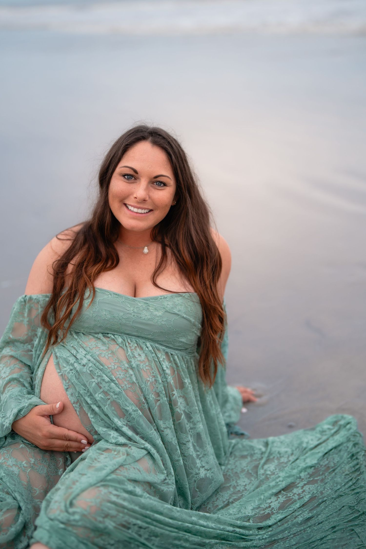 Smiling pregnant woman in teal lace dress sitting on beach shore cradling baby bump during maternity photo session.