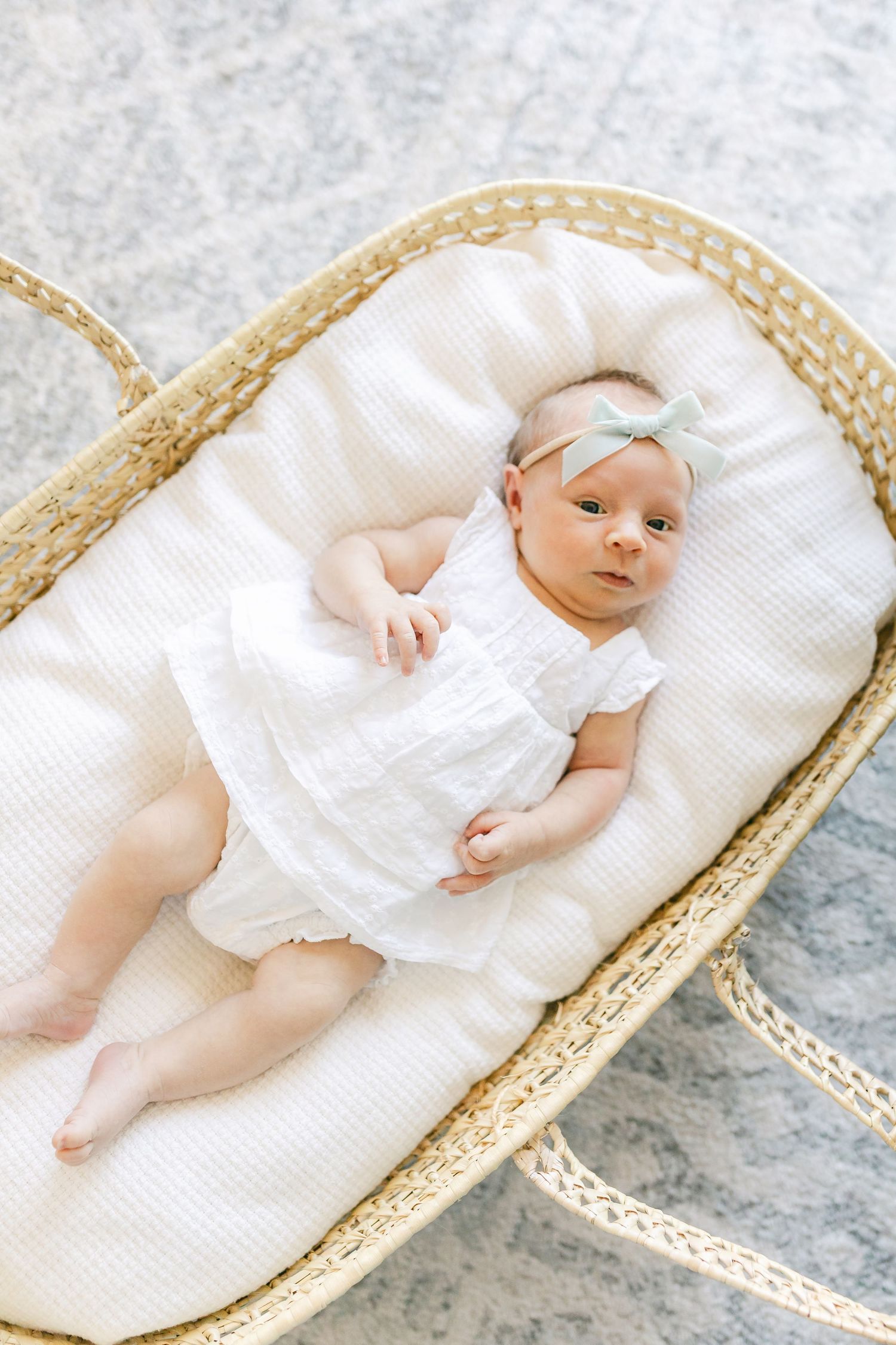Newborn baby girl in white romper with mint bow headband lying in a wicker Moses basket on a soft rug.