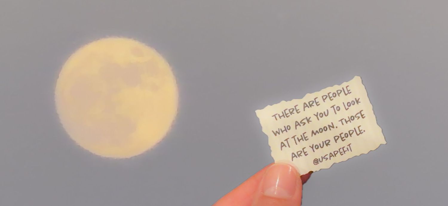 Hand holding a note about moon-gazing friends in front of a full moon in a cloudy sky.
