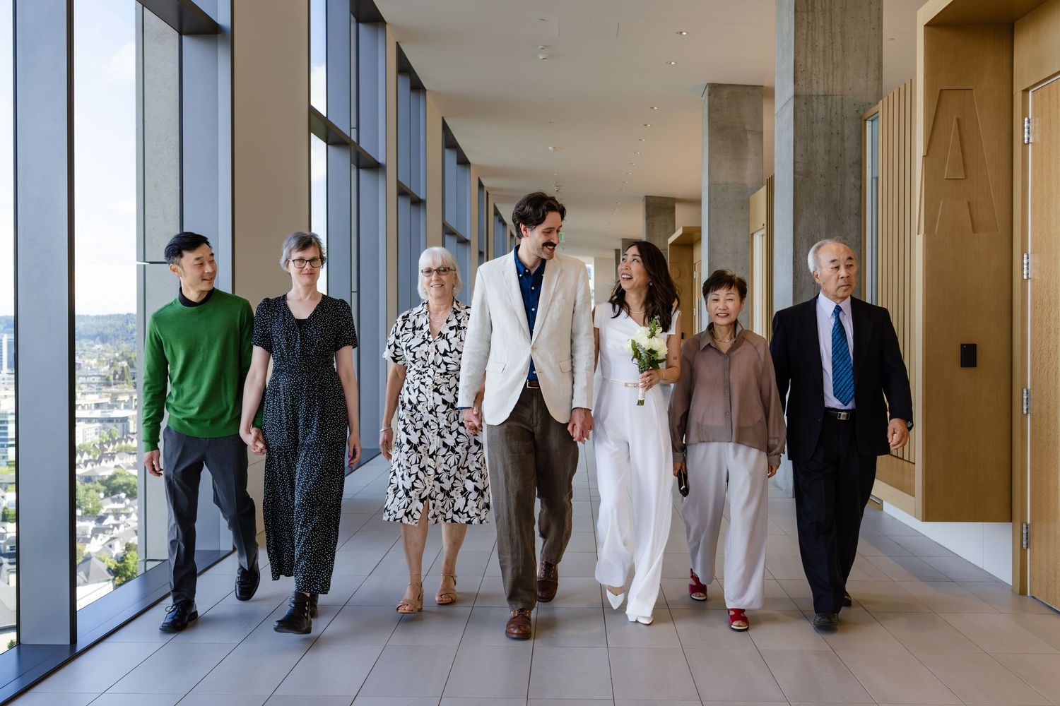 Wedding party walks down a sunlit modern hallway, bride holding white flowers, flanked by smiling family members in formal attire.