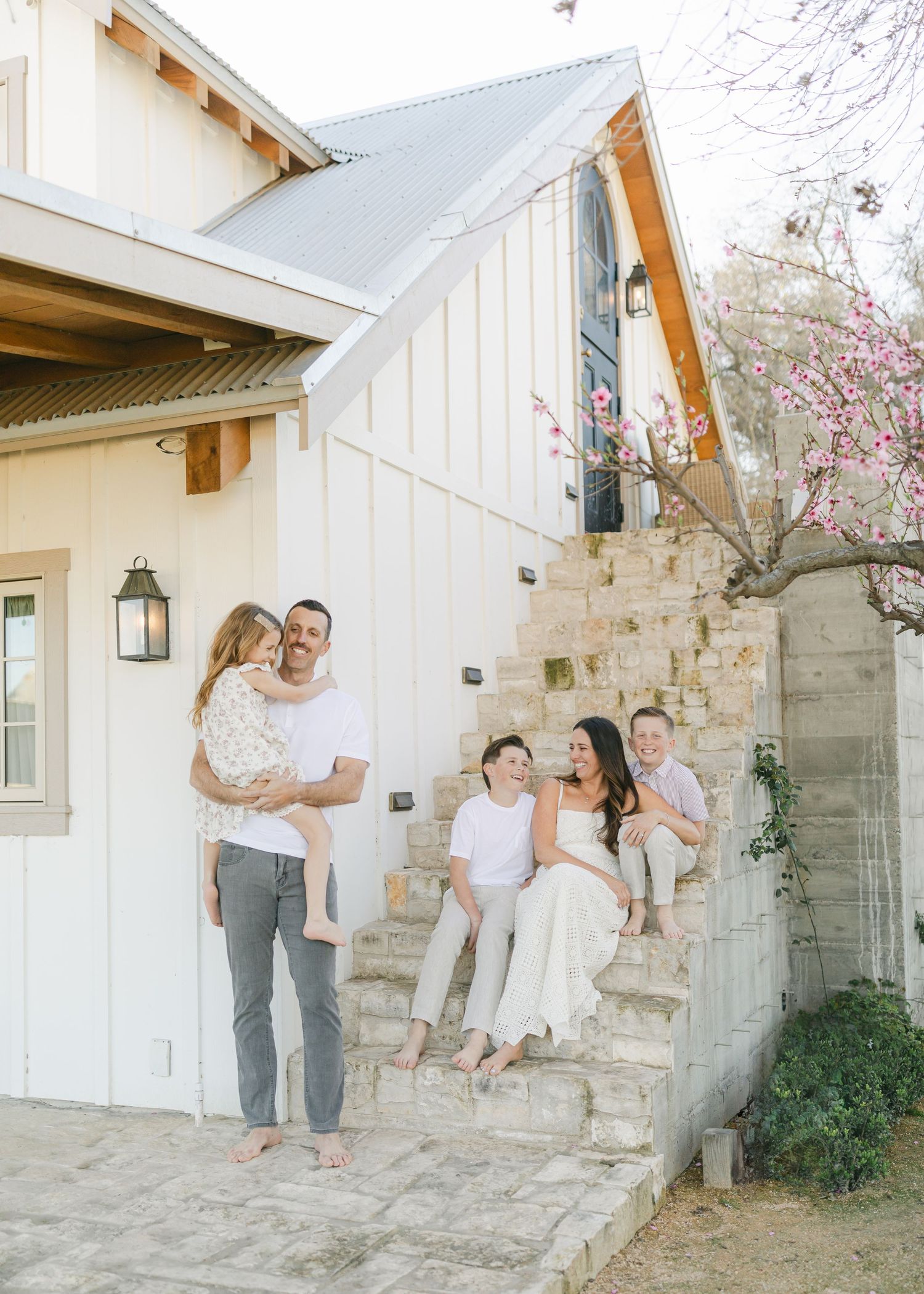 A happy family of five poses on stone steps outside a charming white farmhouse with blooming trees nearby.