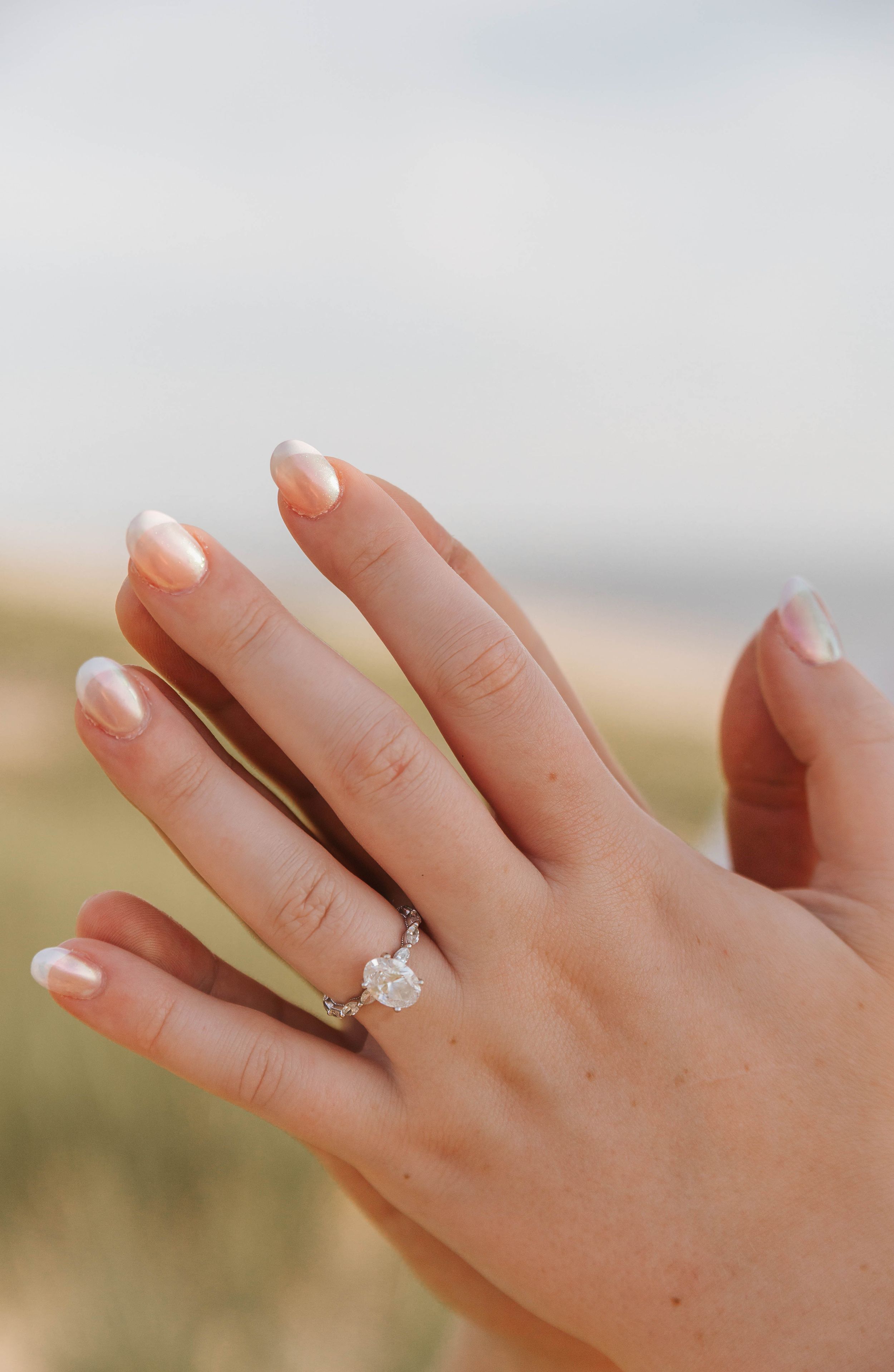 Close-up of a hand with peach nails wearing a stunning solitaire diamond engagement ring on the ring finger.