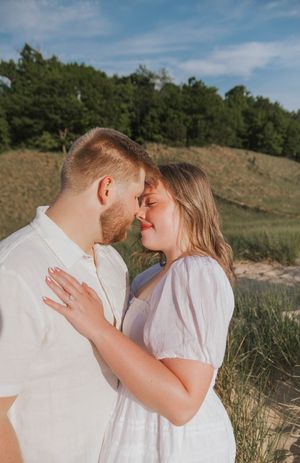 Romantic couple in white outfits sharing an intimate moment outdoors with sandy dunes and green trees in background.