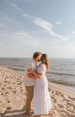 A couple shares a romantic kiss on a sandy beach with calm ocean waves and a blue sky behind them.