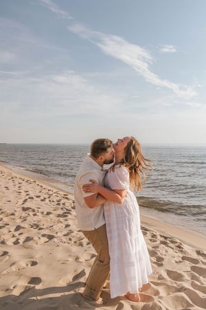 A couple embraces and laughs on a sandy beach, the woman in a white dress, with calm ocean waters behind them.