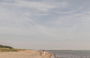 Couple walking hand in hand along a serene sandy beach shore under a vast cloudy sky.