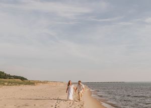 A couple walks hand-in-hand with a child on a sandy beach near calm waters under a partly cloudy sky.