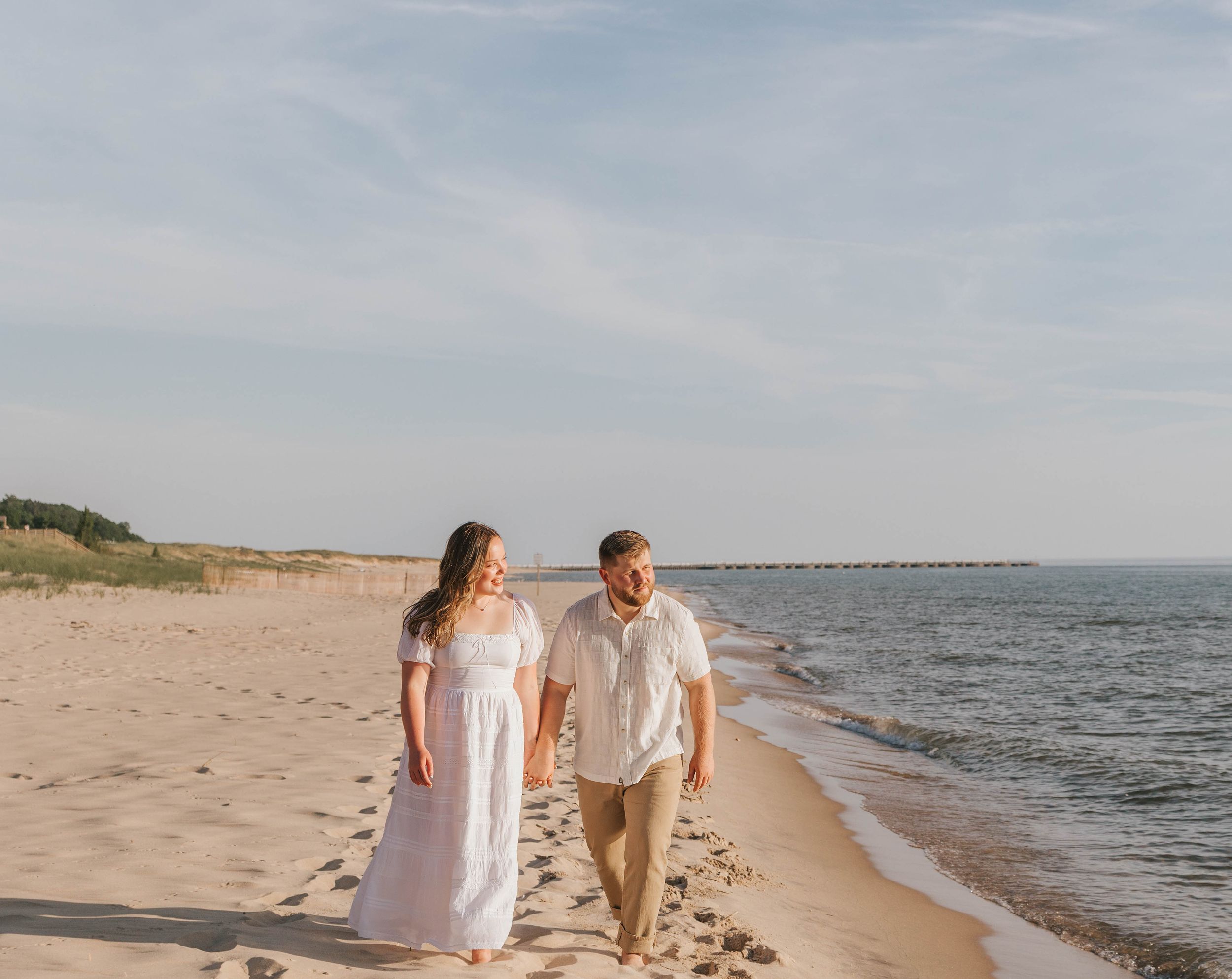 A couple walks hand-in-hand along a sandy beach shoreline, woman in white dress, man in casual attire, on a sunny day.