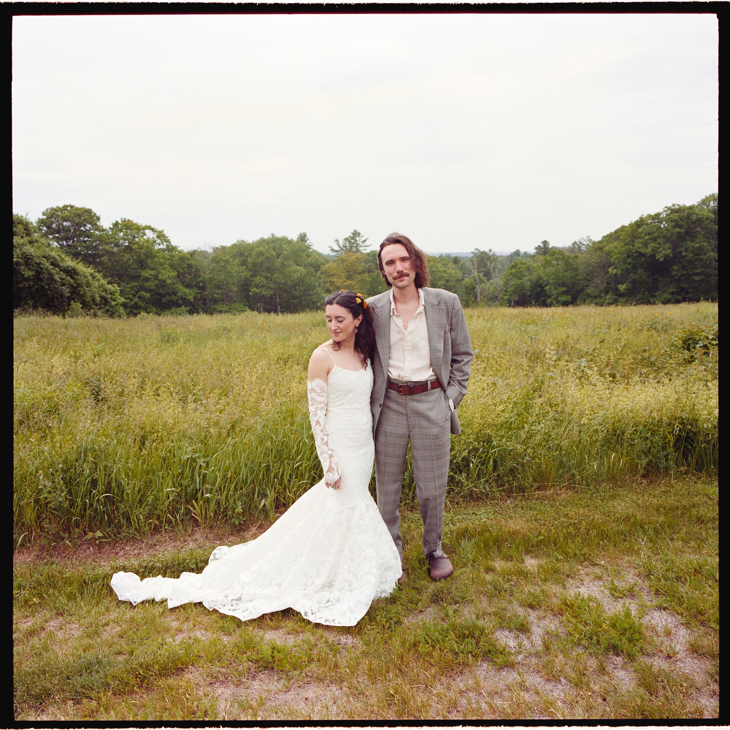 Bride in white lace gown and groom in gray suit pose together in a lush green meadow on their wedding day.