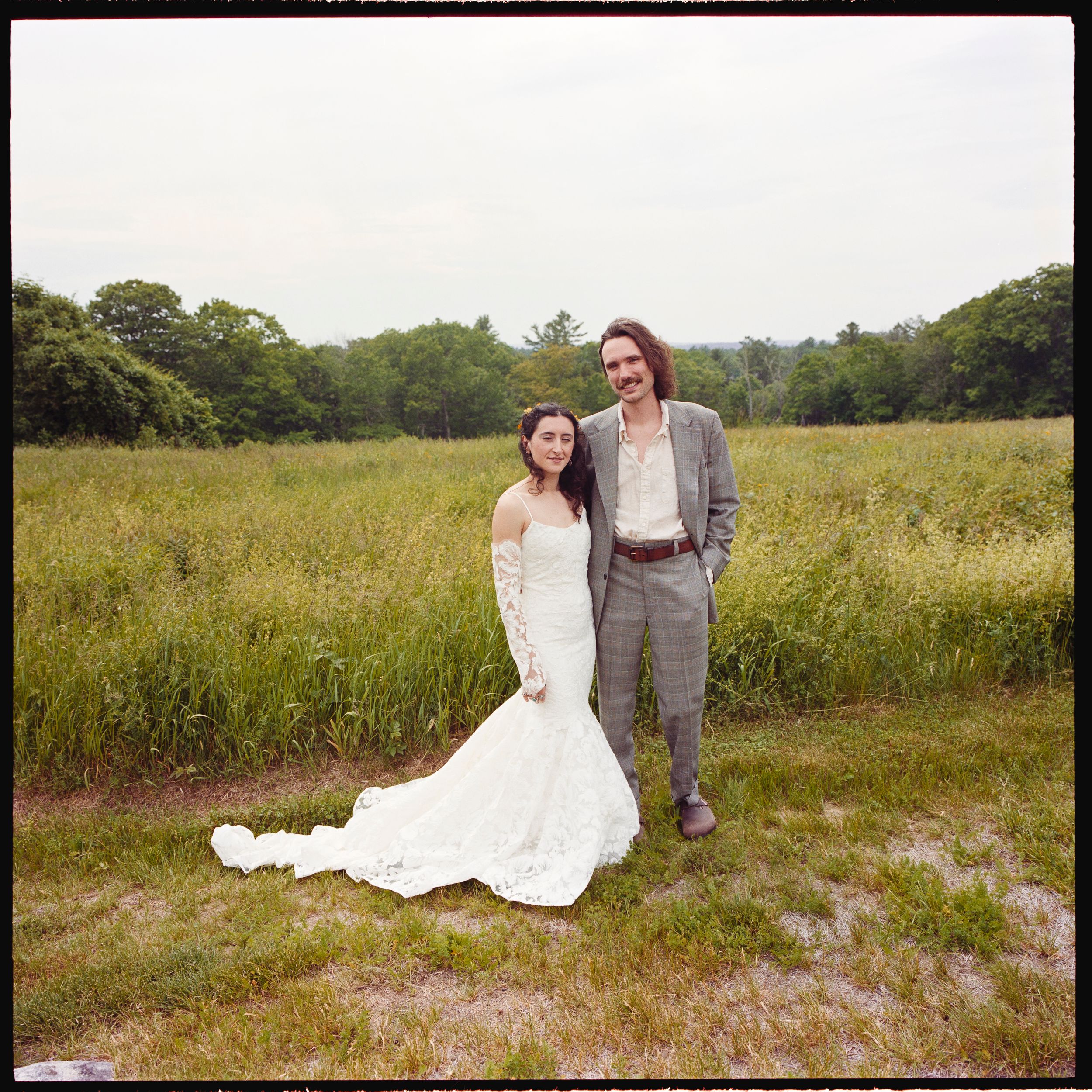 Bride in white lace gown and groom in gray suit pose together in a lush green meadow on their wedding day.