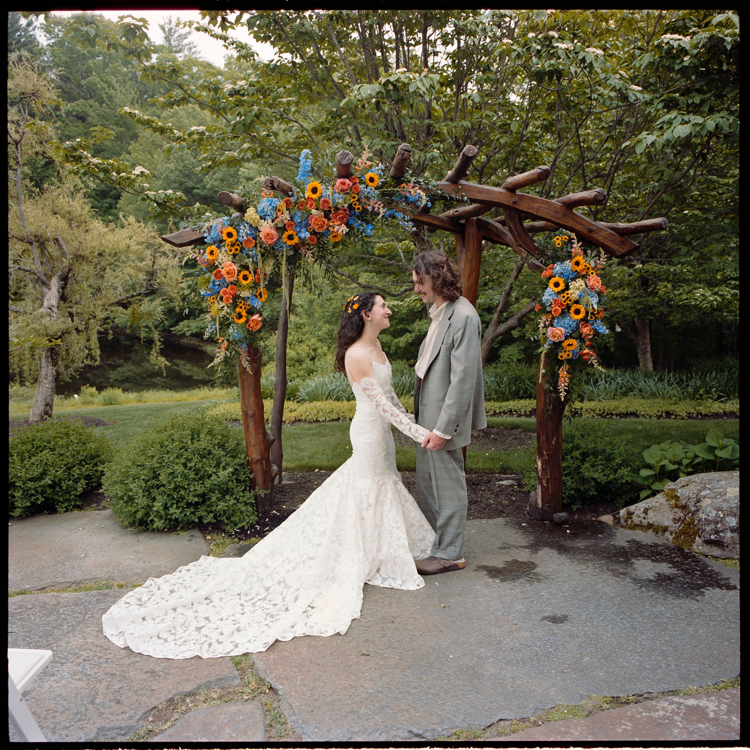 Bride and groom hold hands under a rustic wooden arch adorned with colorful orange and blue flowers in a lush garden.