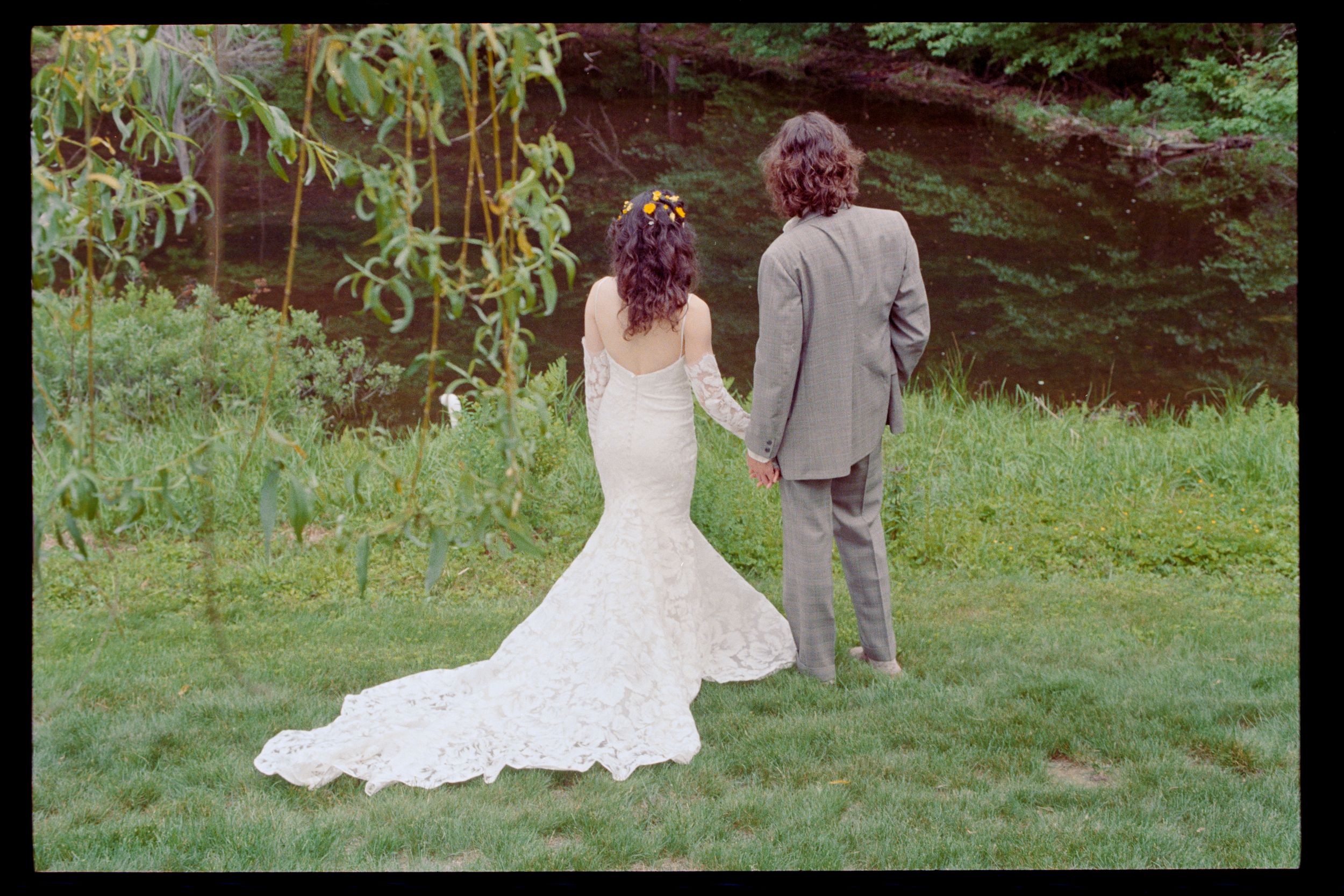 A bride in a lace mermaid gown and groom in gray suit hold hands facing a serene green woodland backdrop.
