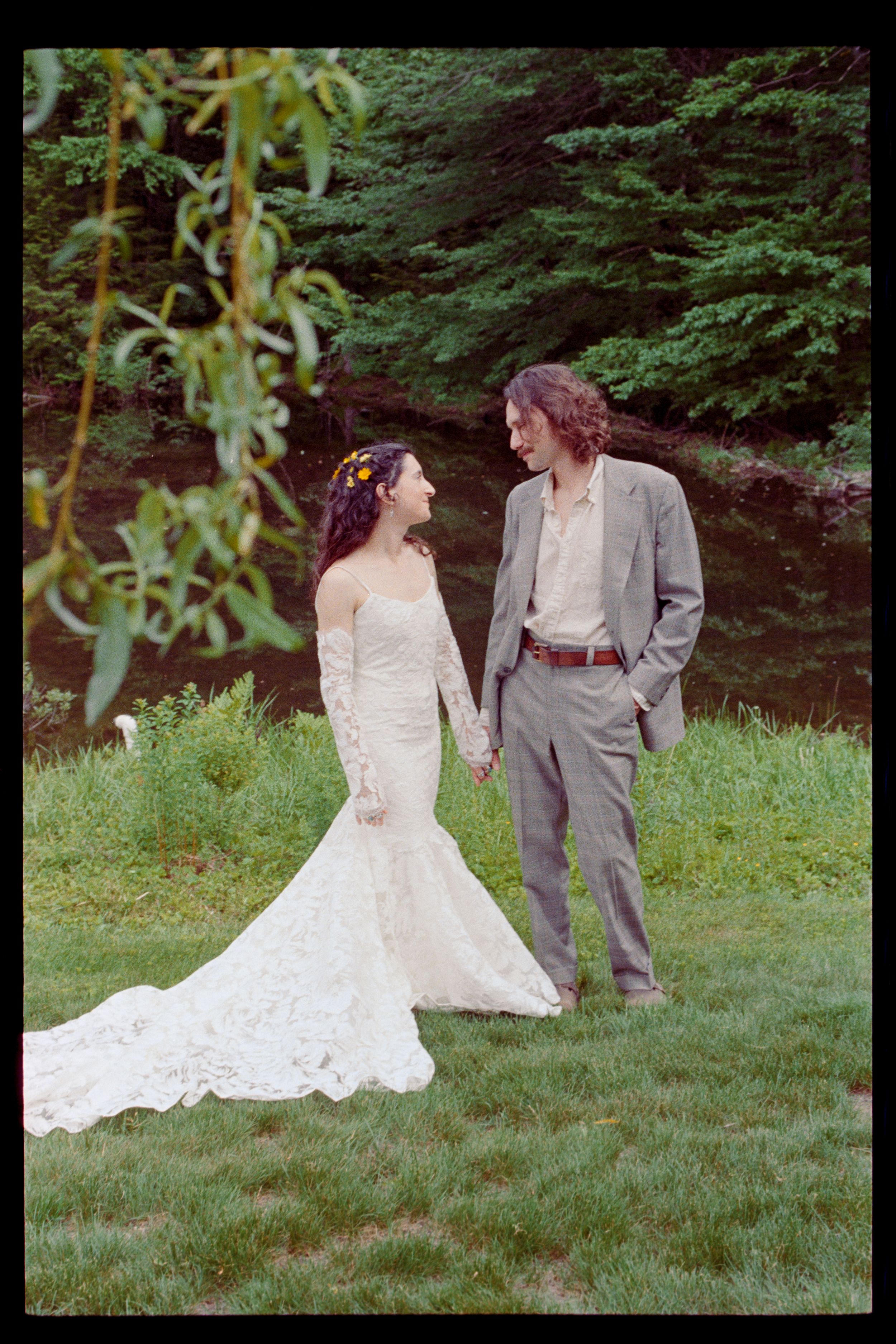 Bride in lace mermaid gown and groom in gray suit hold hands outdoors near a creek surrounded by lush greenery.