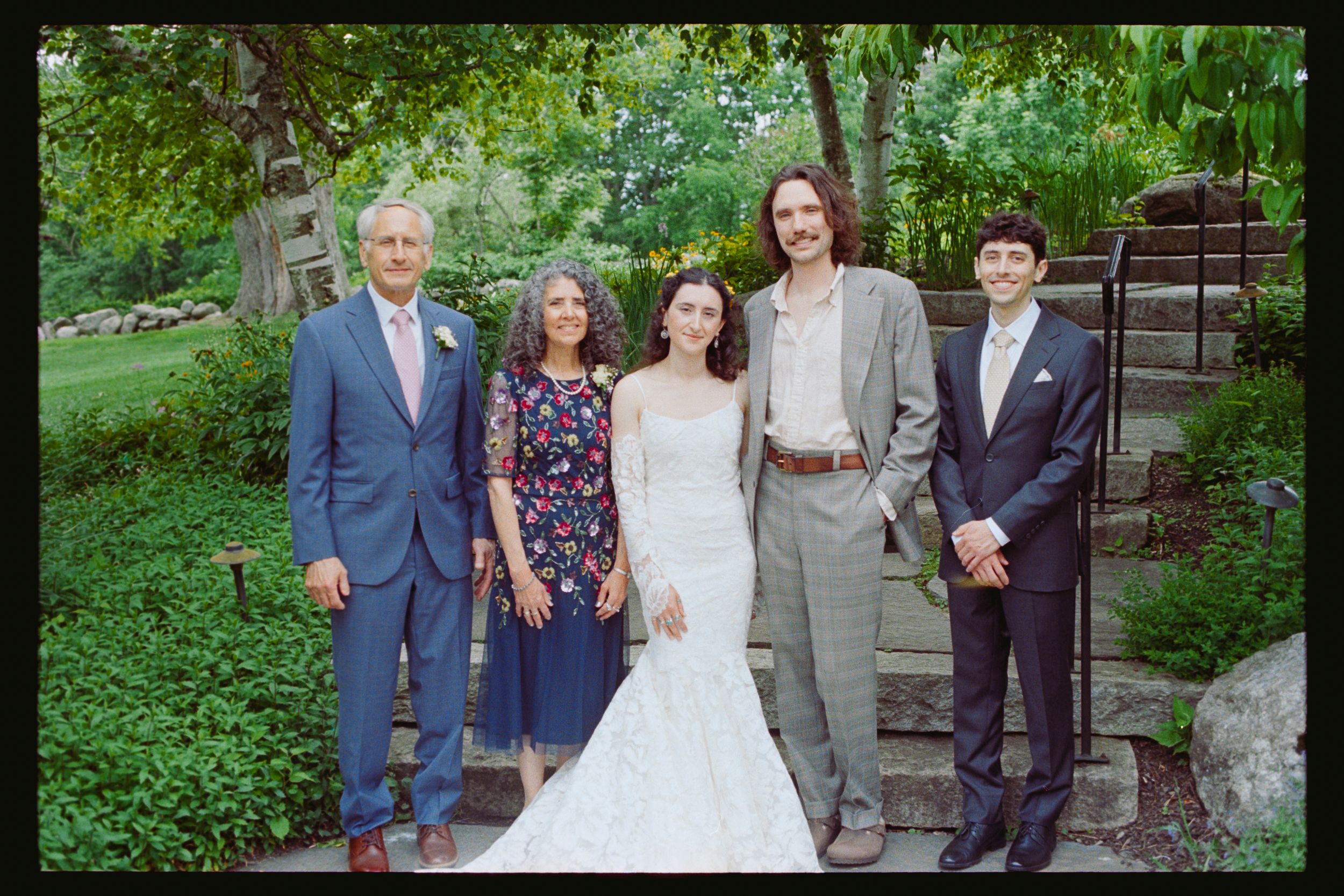 A joyful wedding party poses outdoors, featuring a bride in a white gown with family members dressed in formal attire.