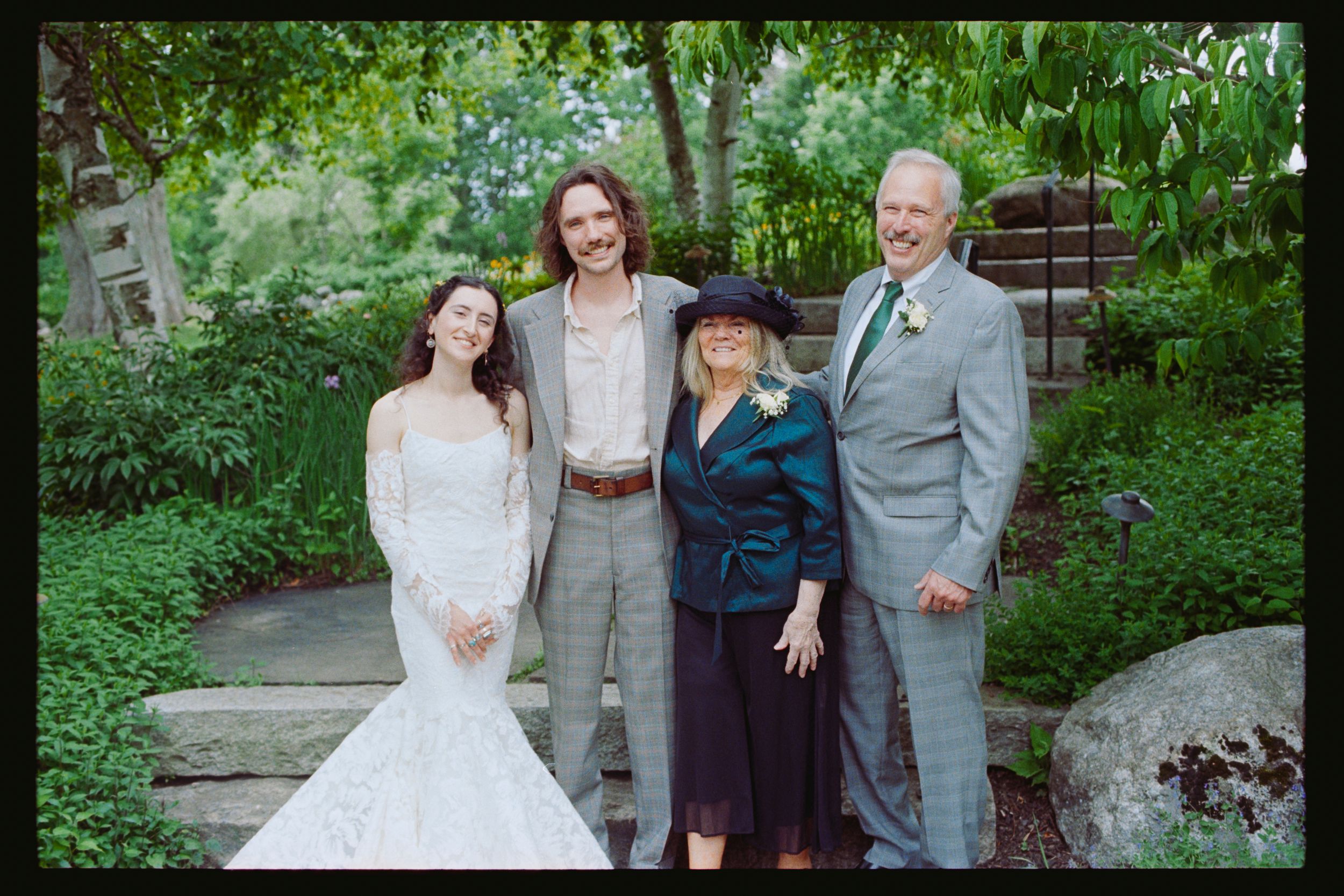Wedding party of four posing in lush garden, bride in white lace gown with groom and elegantly dressed family members.