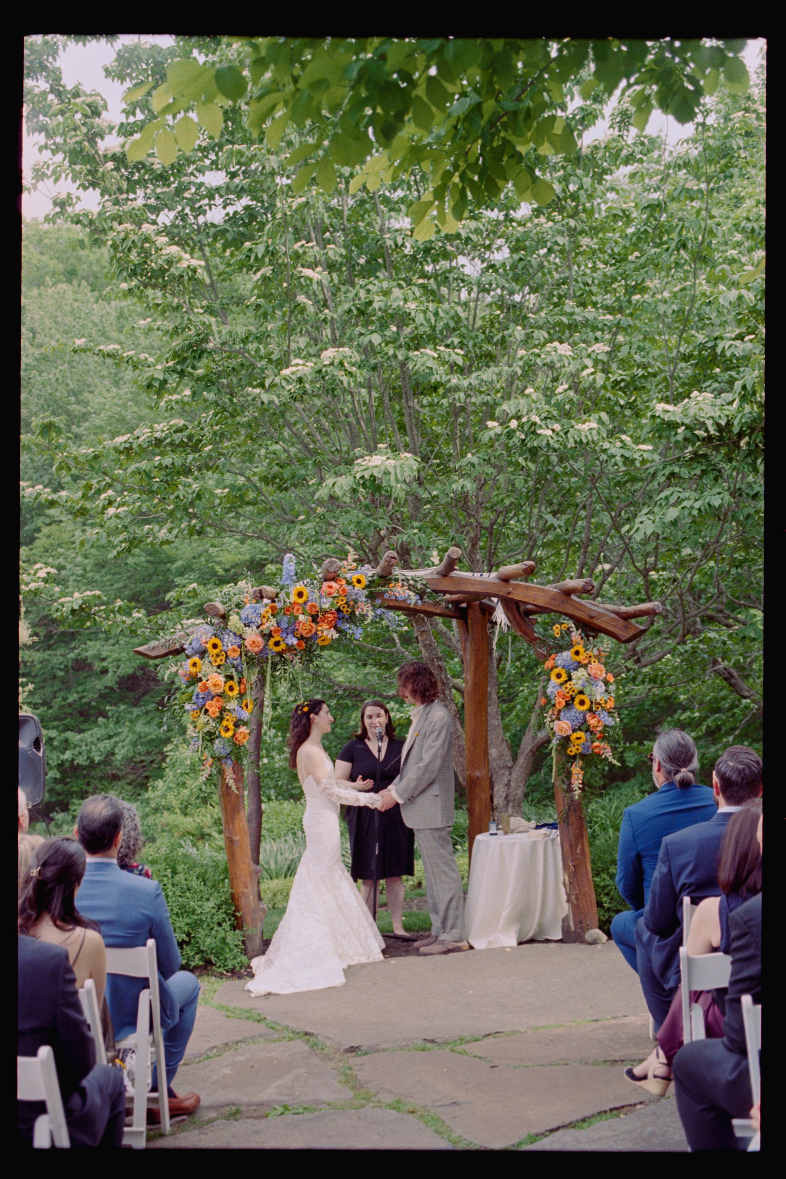 Outdoor wedding ceremony under rustic wooden arch adorned with sunflowers, couple exchanging vows before seated guests.