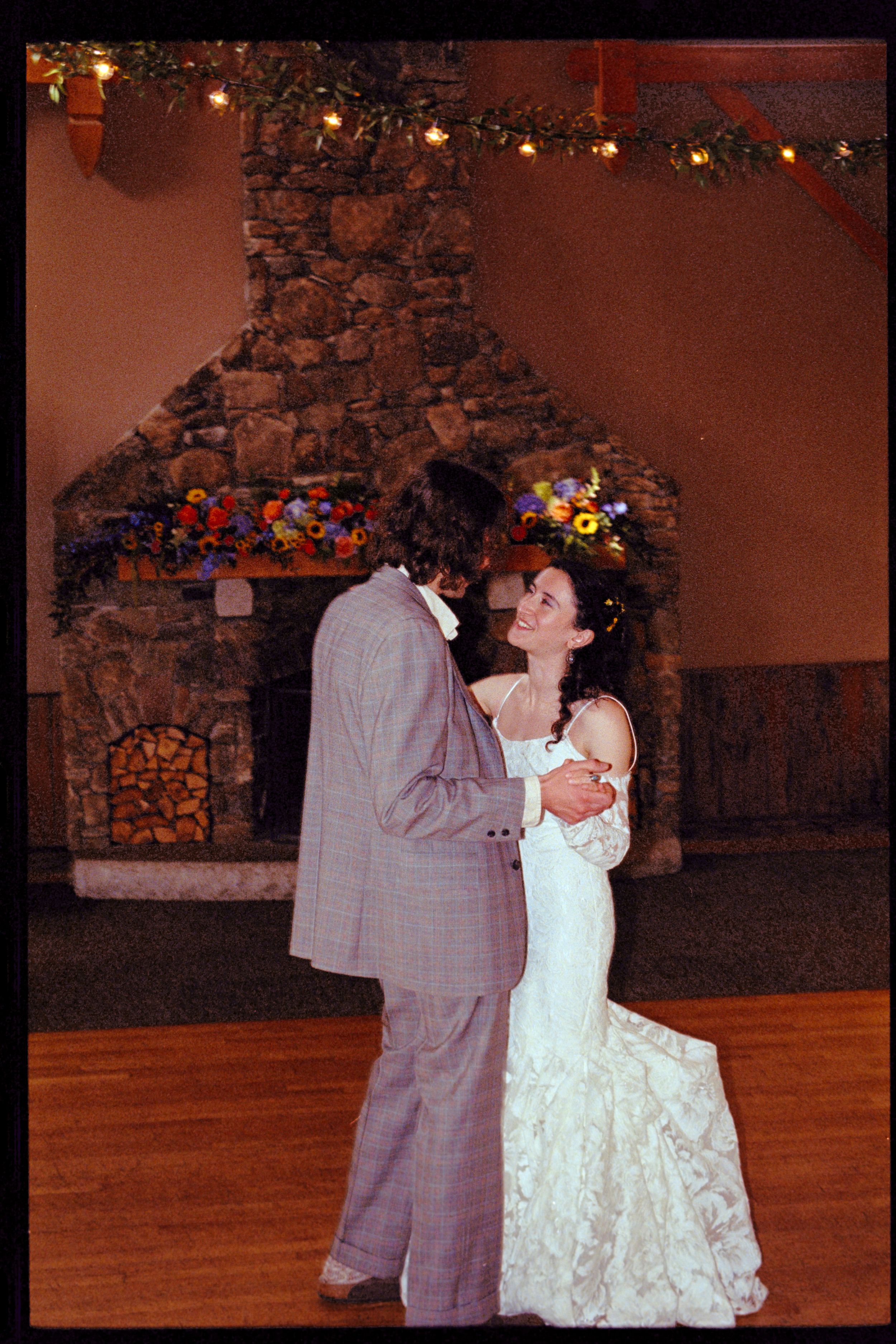Bride and groom share first dance near stone fireplace decorated with colorful flowers at wedding reception.