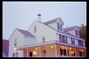 Large white two-story farmhouse with wrap-around porch, multiple dormers, and chimney photographed at dusk.
