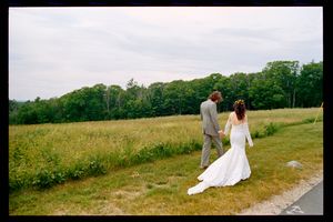 Bride and groom walking hand in hand through a scenic meadow with lush green trees on their wedding day.