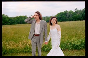 Bride in lace mermaid gown and groom in gray suit hold hands walking through green meadow.