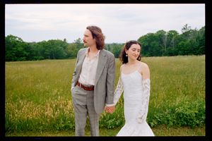 A bride and groom hold hands walking through a lush green meadow, she in a white mermaid gown, he in a gray suit.