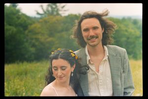 Couple smiling in a sunny field, woman with yellow flowers in hair, man with curly hair and mustache wearing a gray blazer.