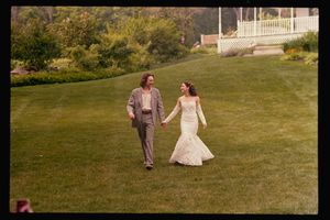 Bride in lace gown and groom in gray suit walk hand-in-hand across a green lawn at an outdoor wedding venue.