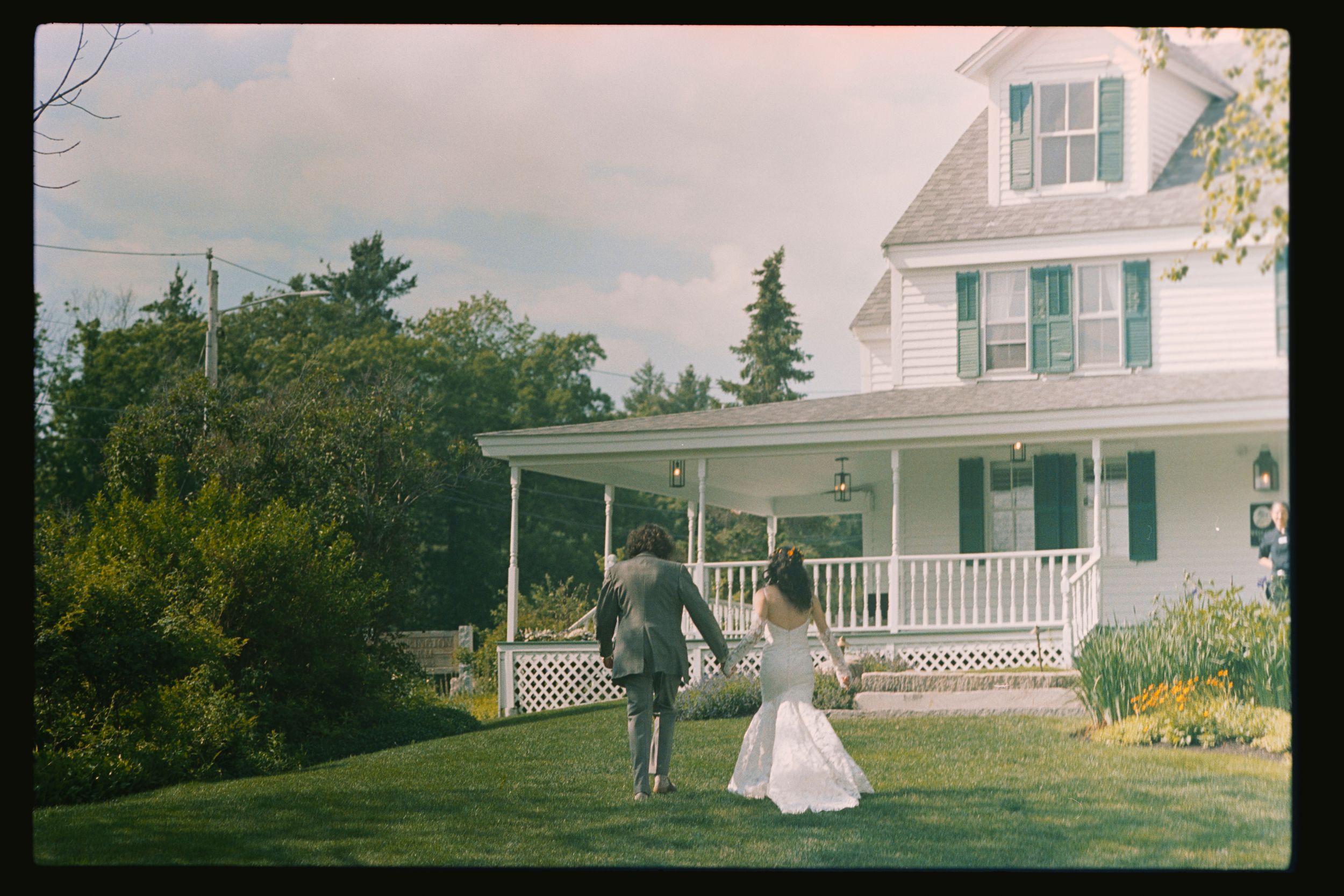 Bride and groom holding hands walking toward a white farmhouse with a wraparound porch on a sunny day.