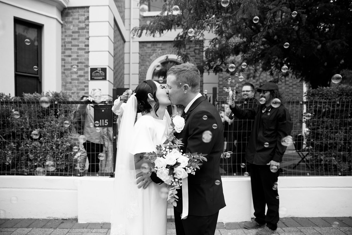 Black and white photo of a bride and groom kissing outdoors surrounded by bubbles and wedding guests.