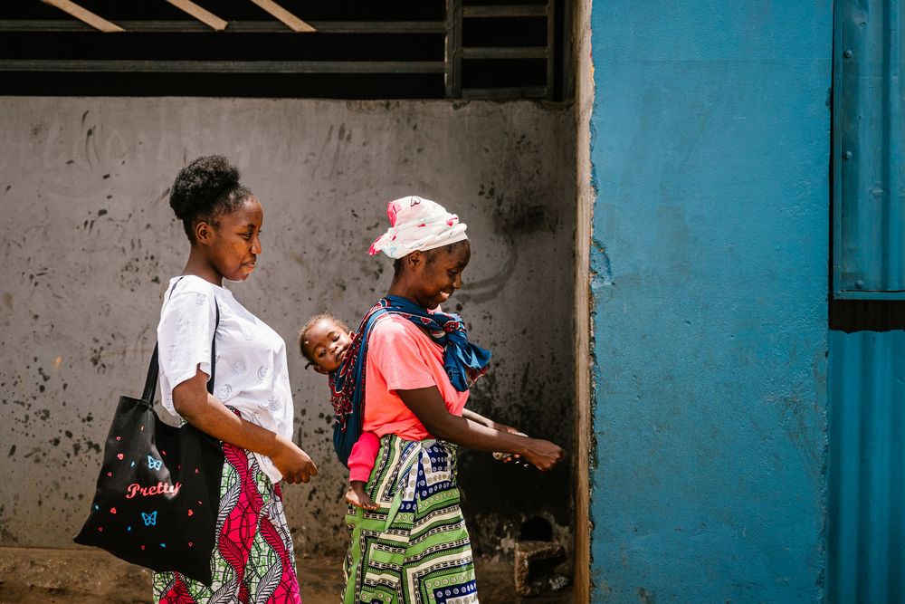 Two African women and a baby walk past a blue door and weathered wall in a vibrant urban street scene.
