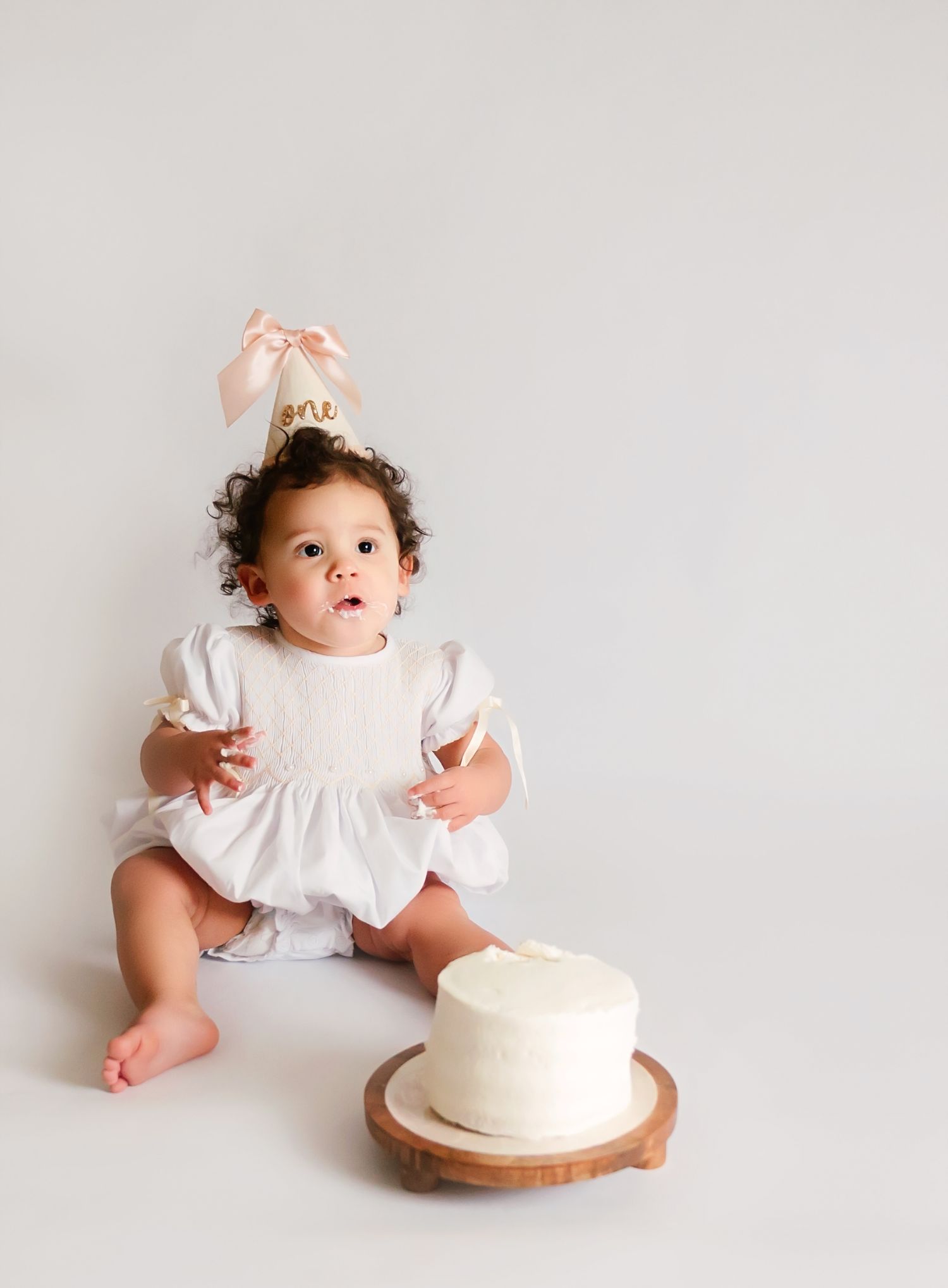 Baby girl in white dress with pink birthday hat sits next to a white smash cake for her first birthday.