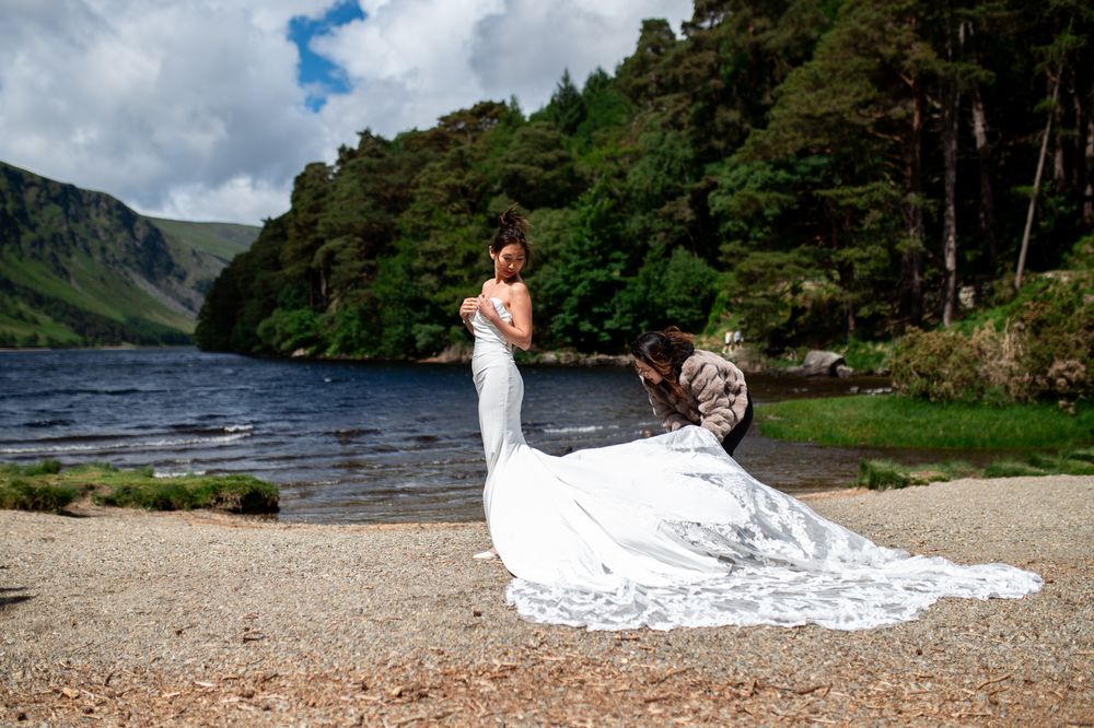 Bride in white gown by a lake with lush green hills, assistant adjusting her train on a pebble shore.