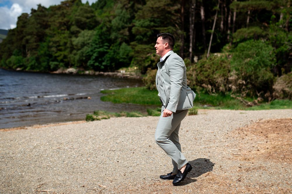 Man in light gray suit walking on sandy shore beside a calm lake surrounded by lush green trees.