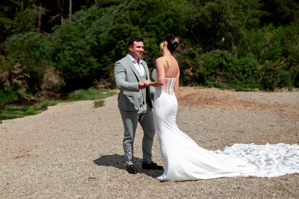 Groom in grey suit shares first look moment with bride in white gown on sunlit gravel path.