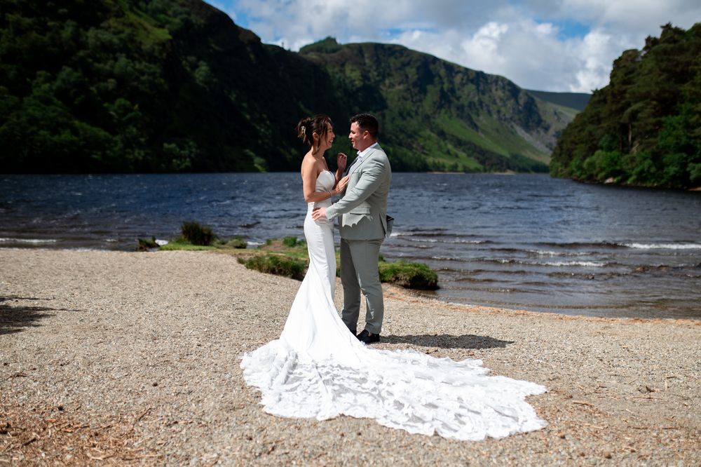 Bride and groom share romantic embrace by serene lake with dramatic green mountain scenery.