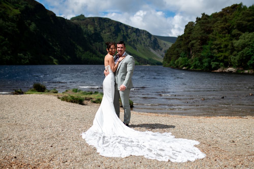 Happy newlyweds pose together on rocky lakeshore with stunning mountain valley background.