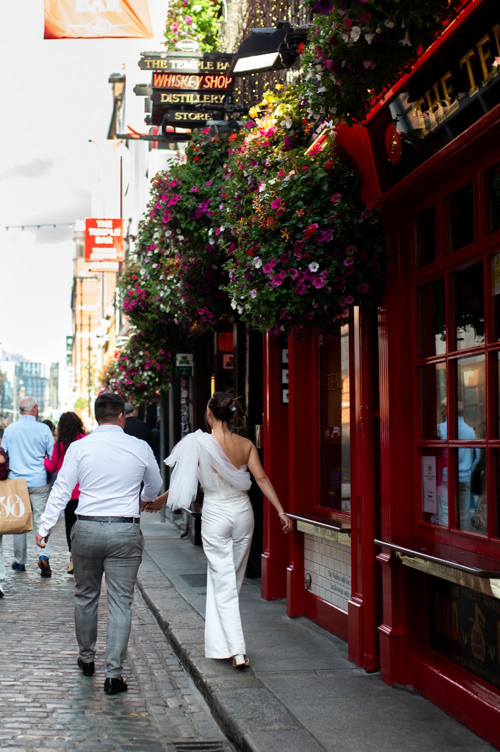Couple holding hands walking past a whiskey shop with hanging flower baskets on a cobblestone street.
