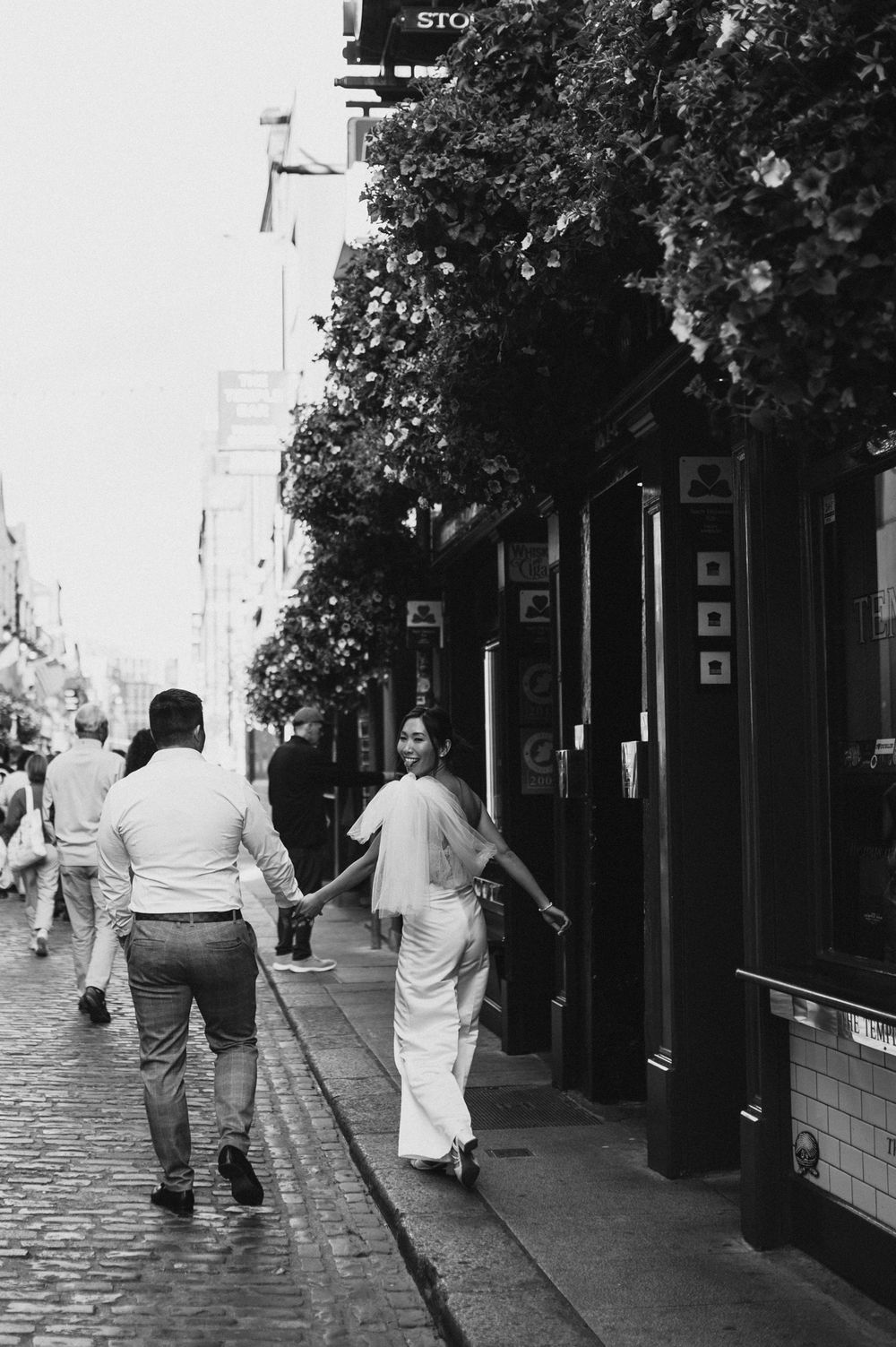 Black and white photo of a couple holding hands walking down a cobblestone street past a pub.