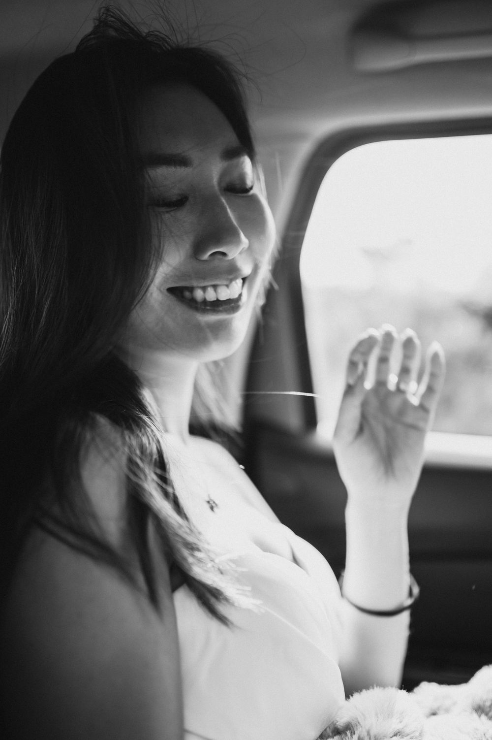 Black and white photo of a smiling young woman with eyes closed sitting in a car, hand raised near face.