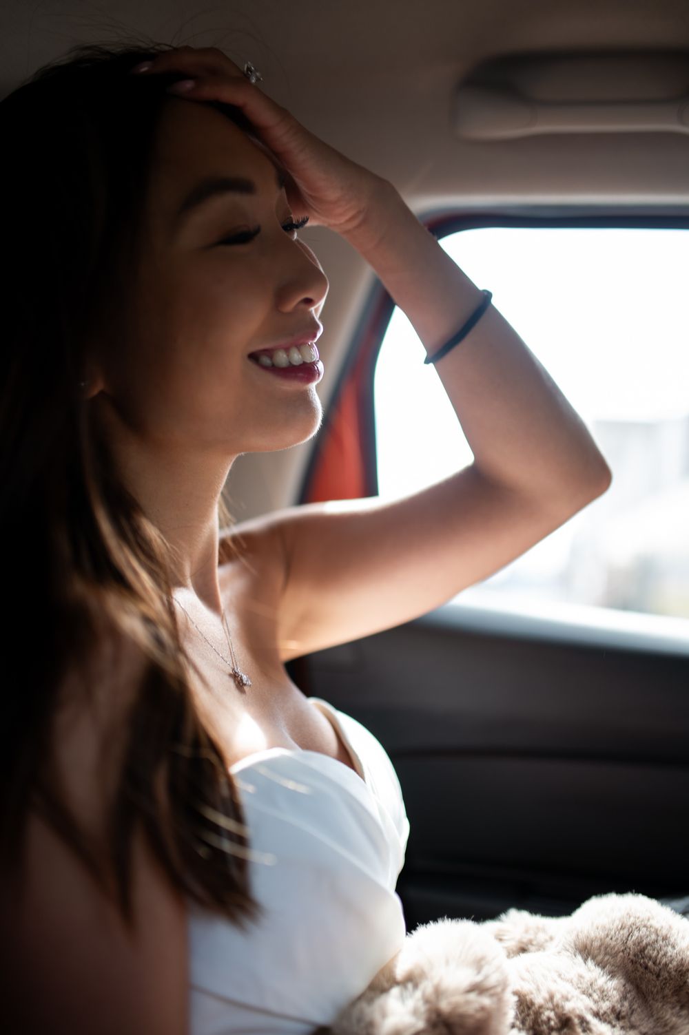 Smiling young woman in white dress sitting in car backseat, hand in hair, sunlight streaming through window.